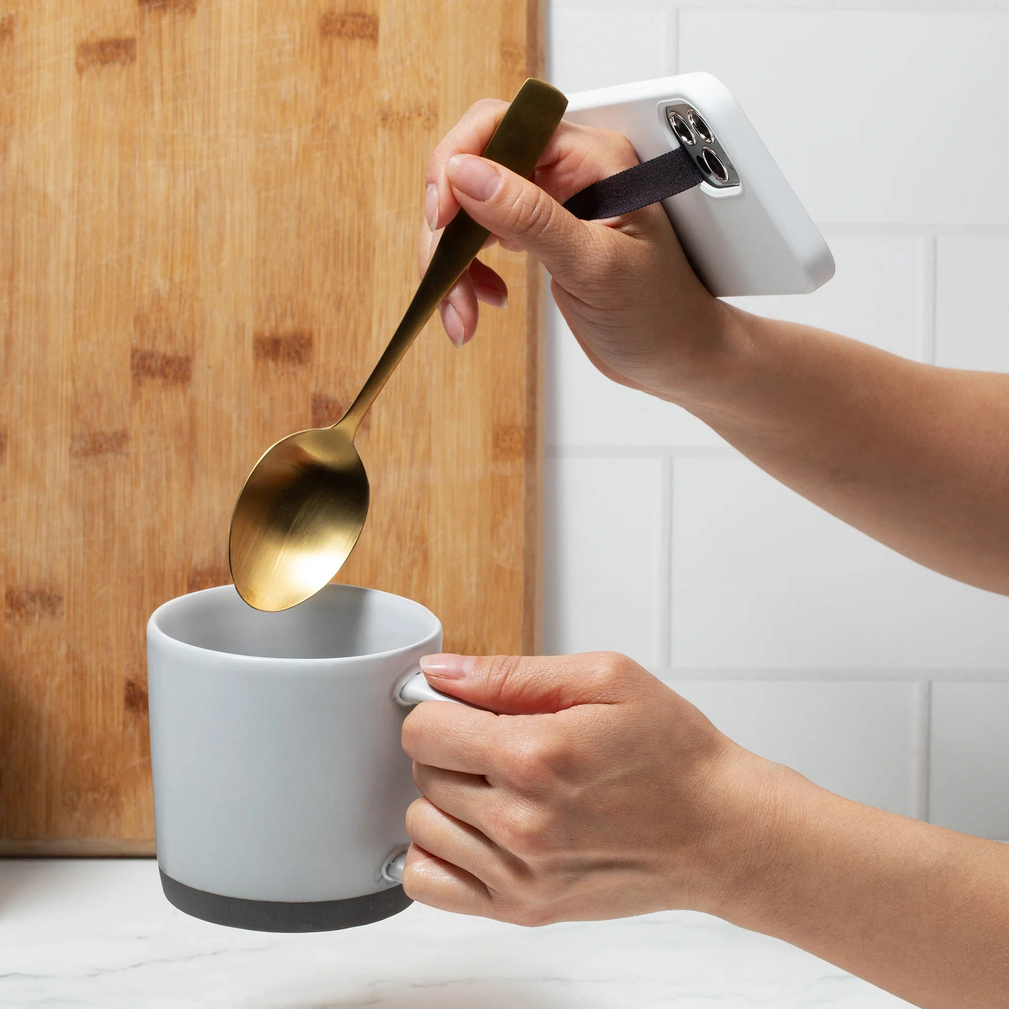 A hand stirring a mug with a spoon while holding a phone by its strap